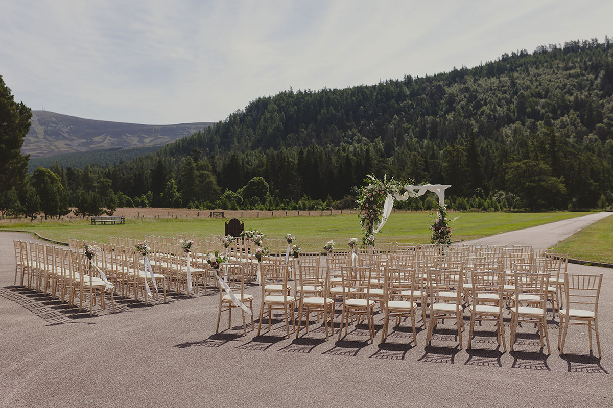 An outdoor wedding ceremony layout with rows of chairs and a floral arch, overlooking open countryside and forested hills.