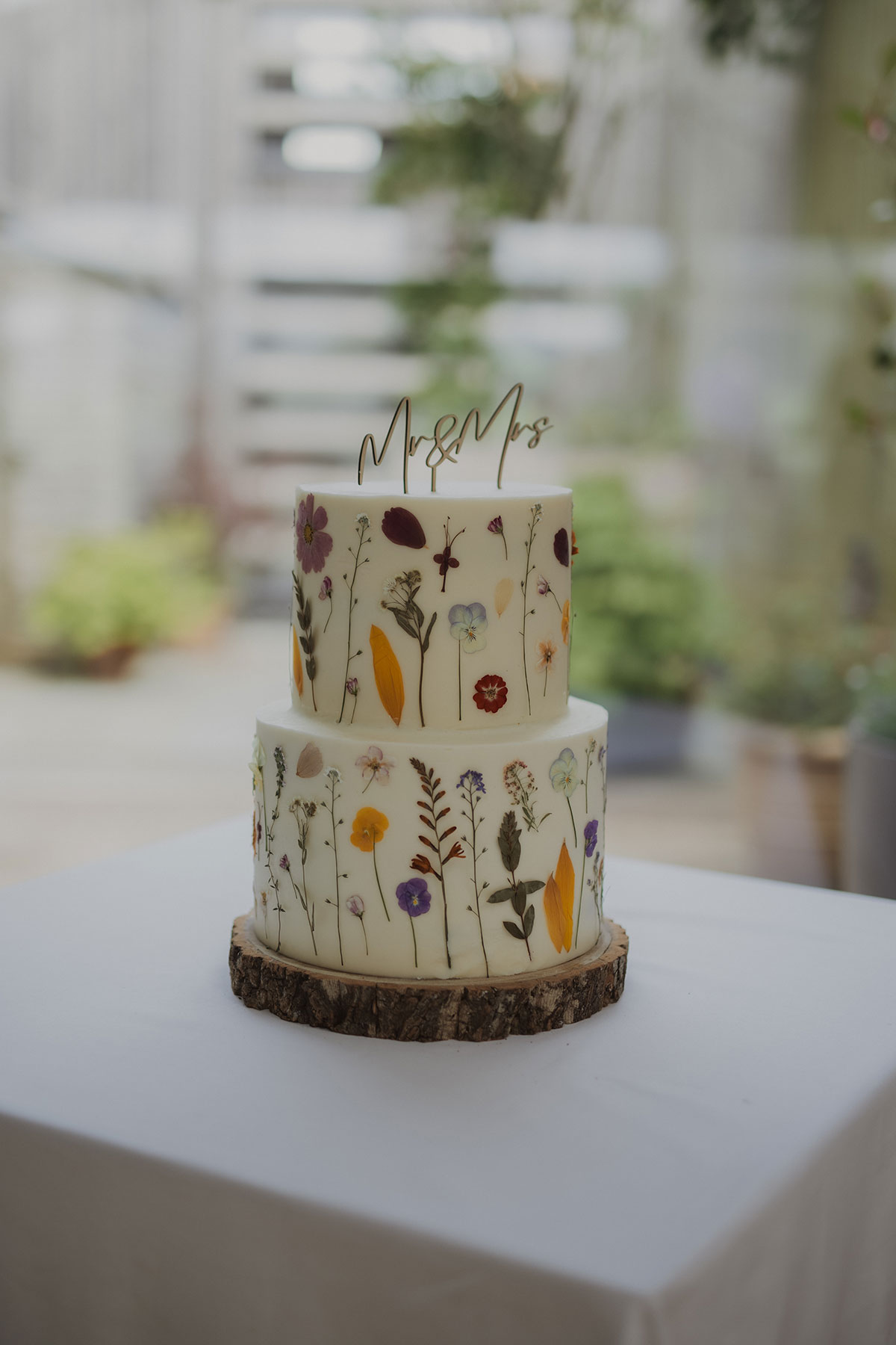 Two-tier pressed flower wedding cake with “Mr & Mrs” topper displayed on rustic wood stand at Scottish reception