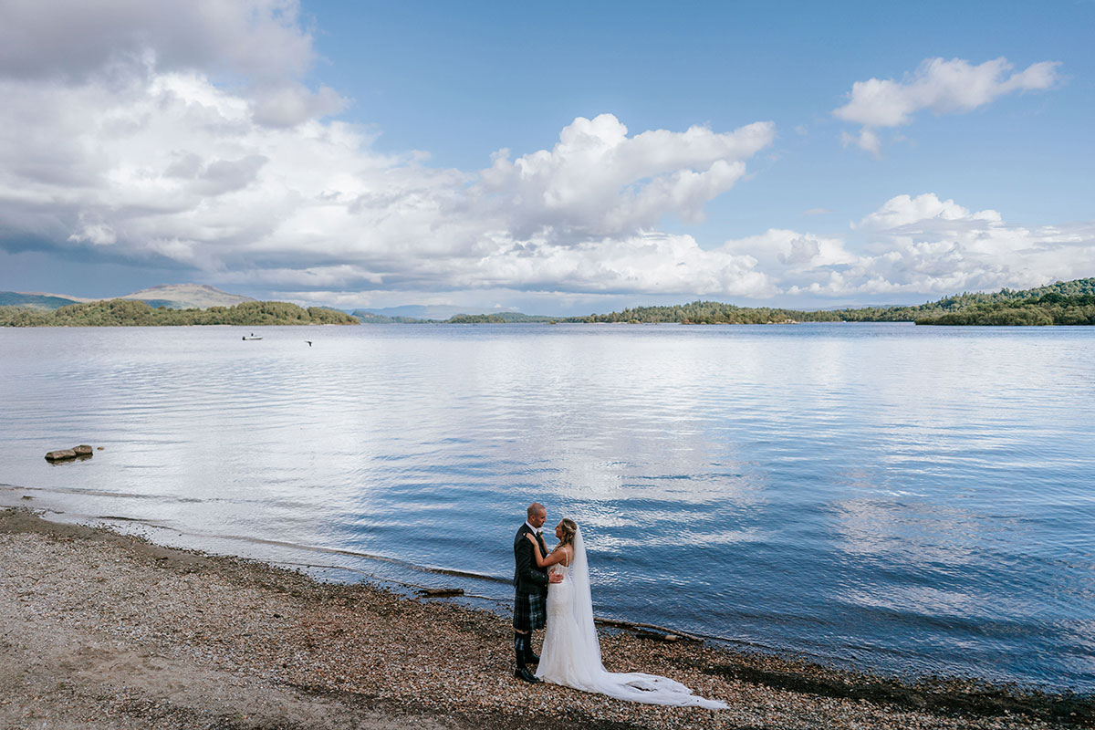 Bride and groom on Loch Lomond shoreline with loch and mountain views at Lodge on Loch Lomond wedding venue