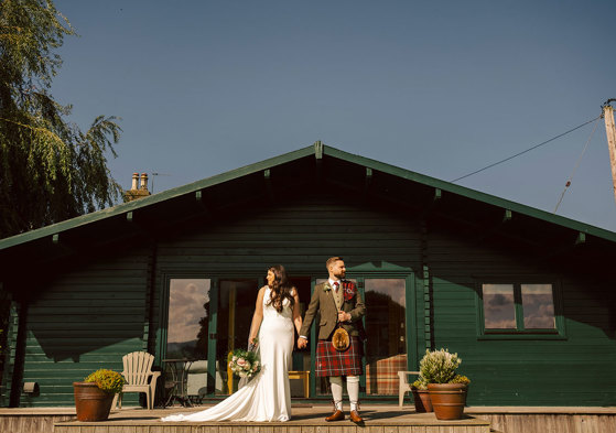 A bride and groom stand in front of a small green building holding hands 