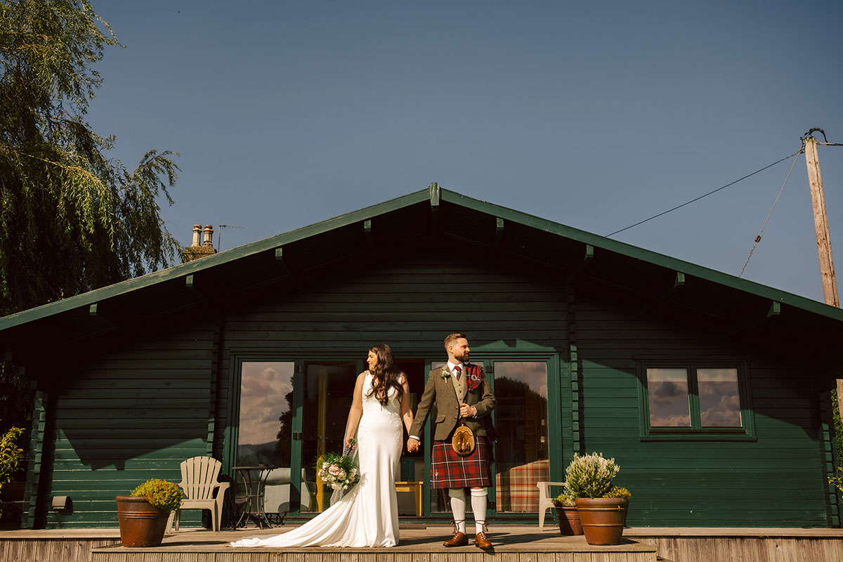 A bride and groom stand in front of a small green building holding hands