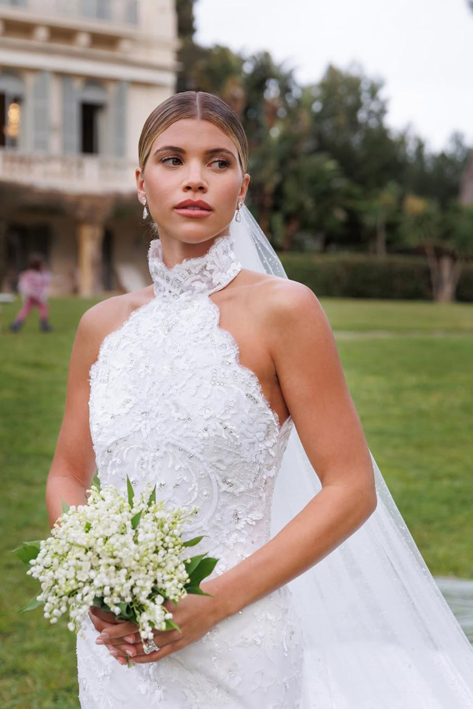 bride wears halterneck embellished wedding dress with a long veil flowing from a low bun and a small bouquet of little white flowers clasped in her hands