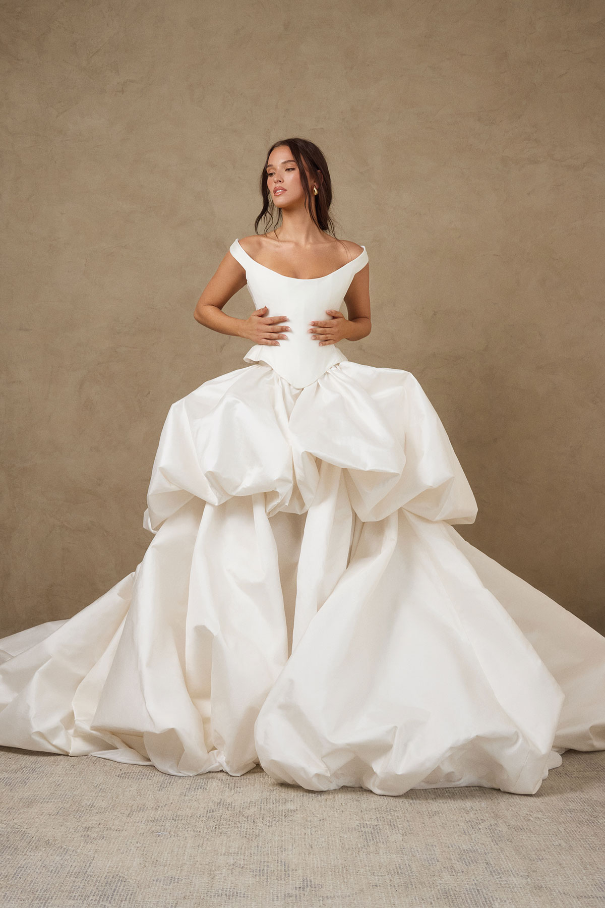brunette model in wedding dress consisting of structured corset and large cloud-like layered ballgown skirt stands with both her arms gently placed on her ribcage, elbows out, face turned to the side