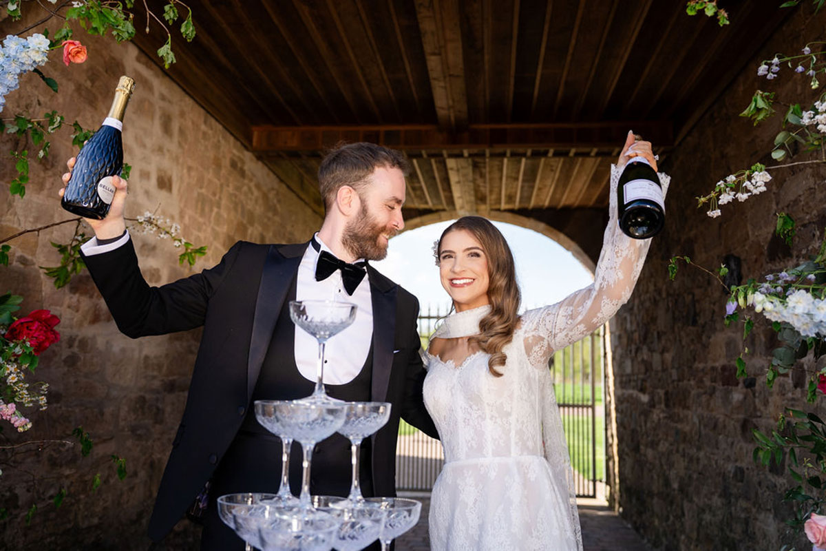 Bride and groom celebrating under a stone archway, each holding a champagne bottle beside a stacked champagne tower decorated with floral arrangements