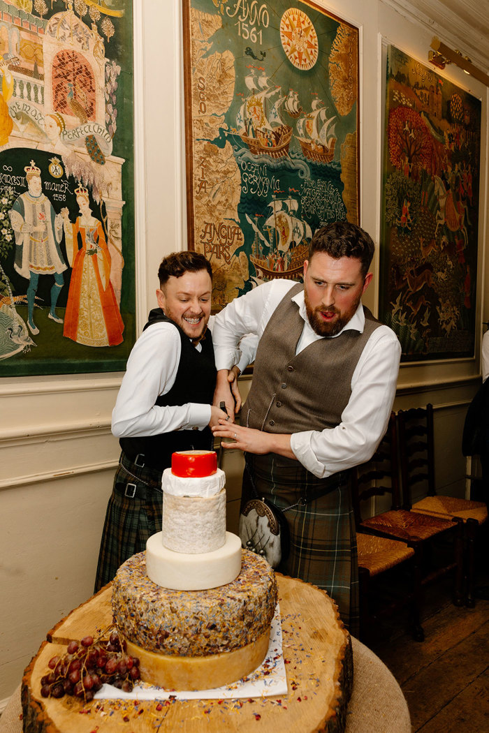 two grooms wearing kilts cutting a tower of cheese 'wedding cake' at their Neidpath Castle wedding