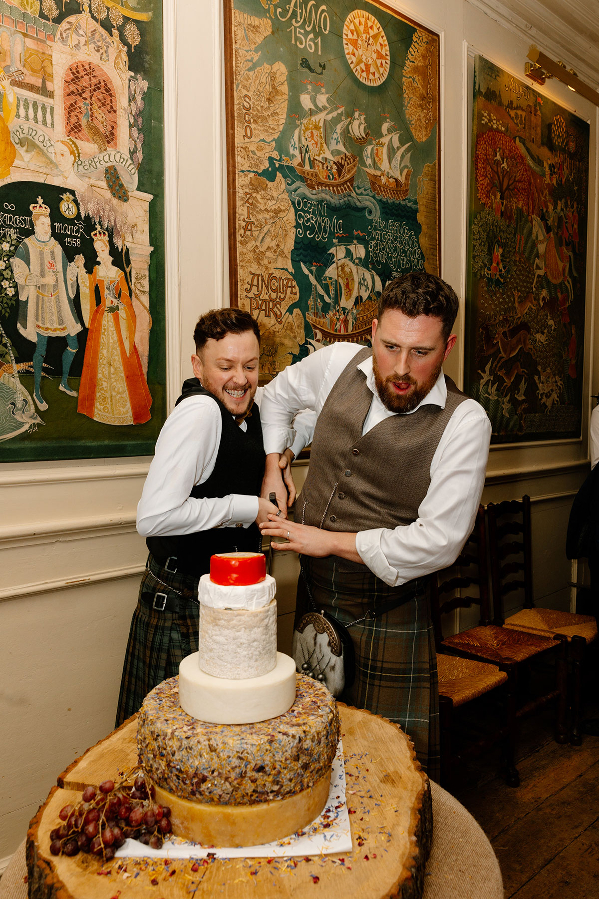 two grooms wearing kilts cutting a tower of cheese 'wedding cake' at their Neidpath Castle wedding