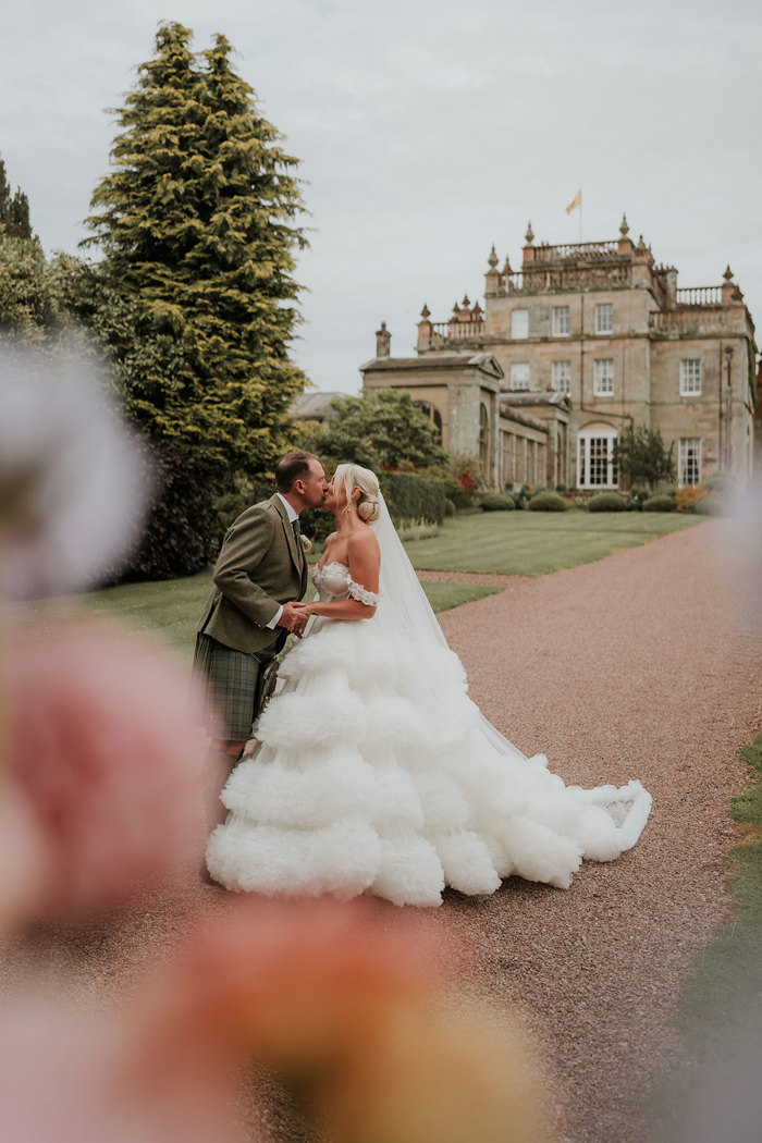 A bride wearing a wedding dress with a voluminous tulle skirt standing next to a groom wearing a green kilt kiss while standing in a garden in front of a country house