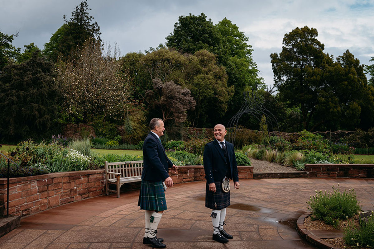 Two grooms in traditional kilts sharing a relaxed moment in the gardens at Brodick Castle before their wedding ceremony.