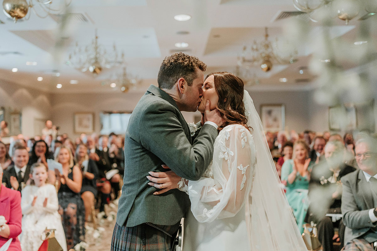 Close-up of bride and groom sharing a kiss during their ceremony, with guests blurred in the background.