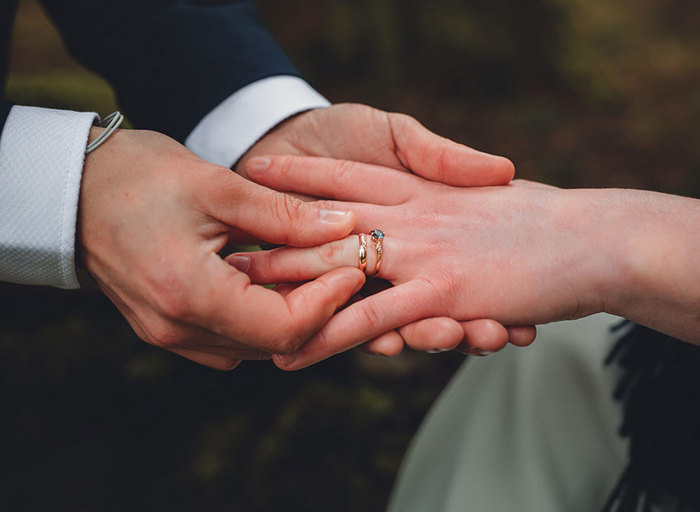 A close up of someone putting a gold wedding band on another person's ring finger