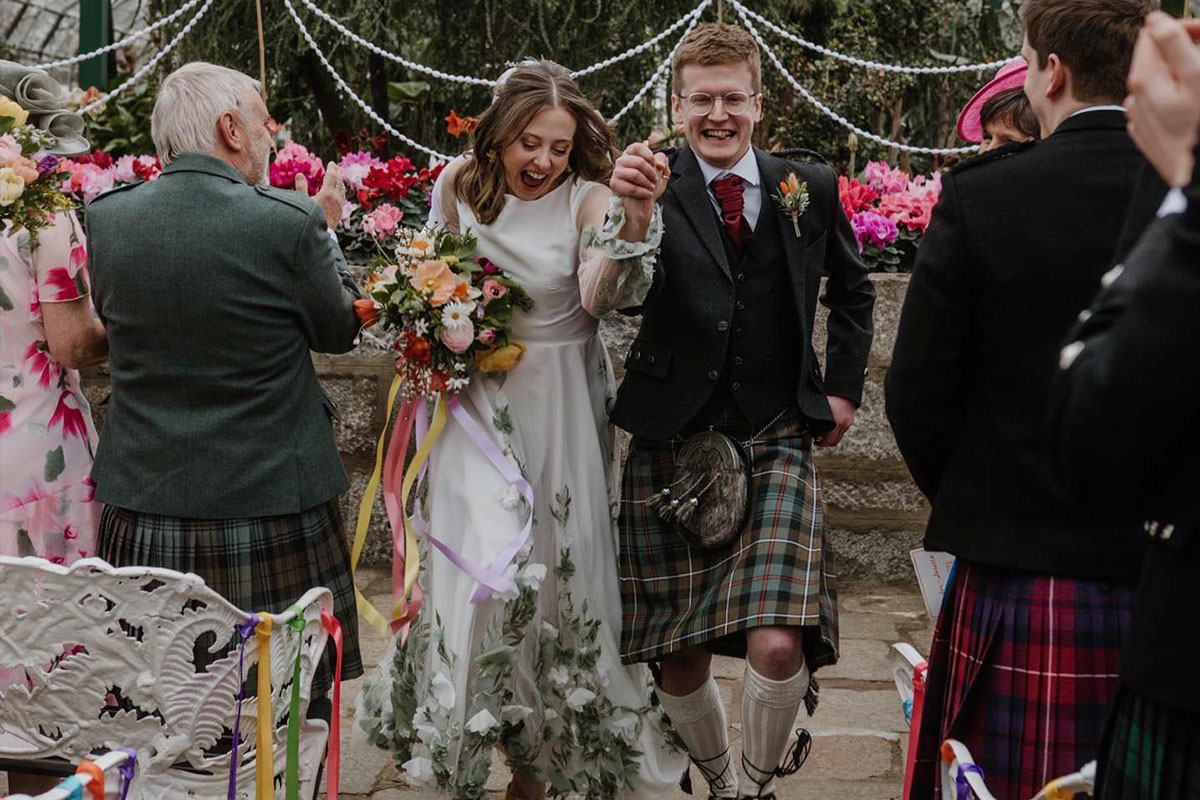 A bride and groom smiling as they walk down the aisle holding hands with their guests on either side clapping