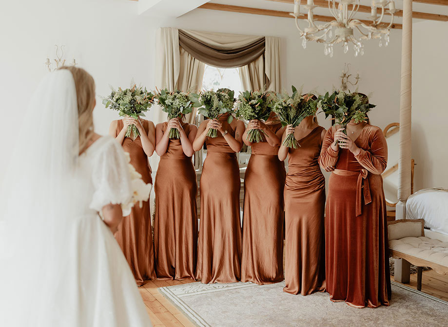 six bridesmaids wearing rust-coloured dresses hold green and white bouquets with orange details up to their faces as bride stands in foreground