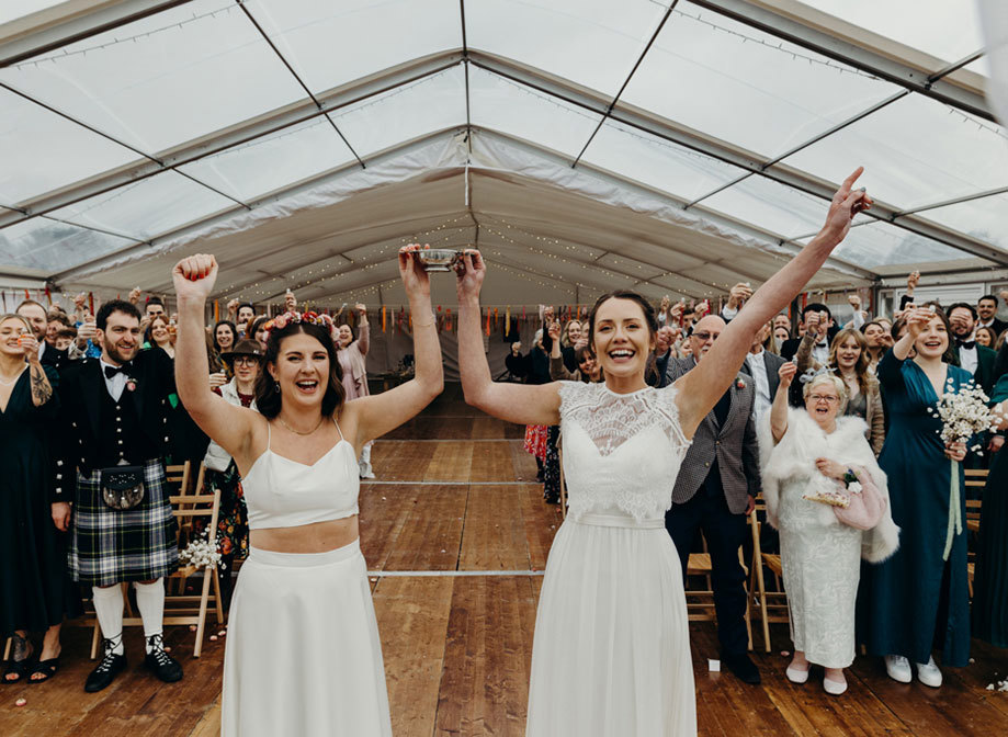 two brides hold a silver quaich aloft and cheer as wedding guests assembled behind them raise small shot glasses and toast. They are in a marquee with wooden floor