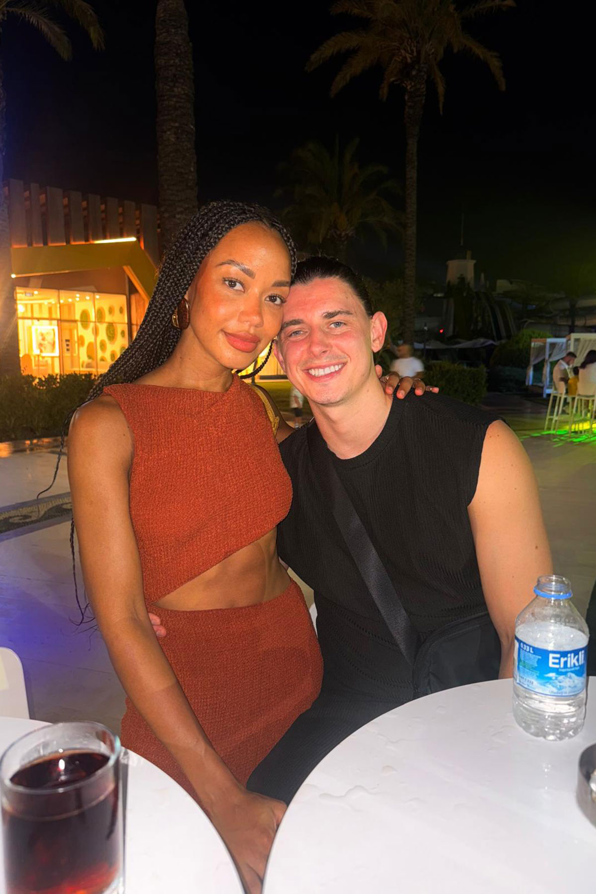 A couple pose together at an outdoor nightlife setting, with palm trees and colourful lights behind them