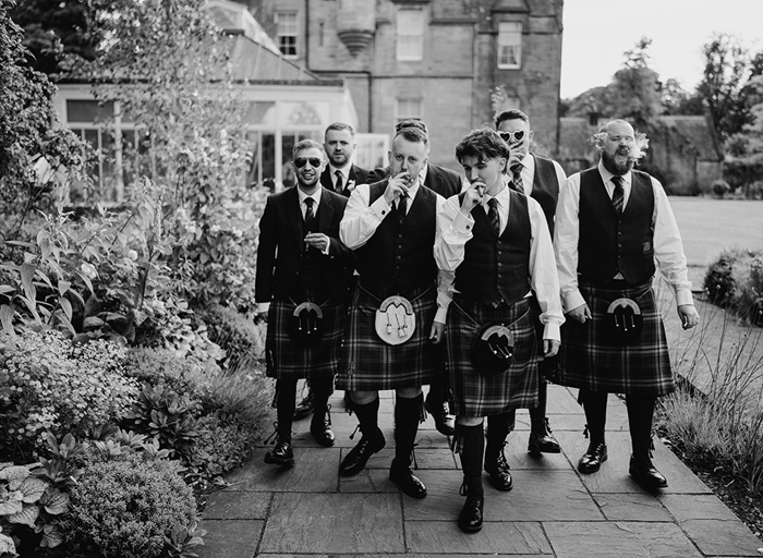 A black and white photo of a group of groomsmen in kilts walking along a path smoking cigars