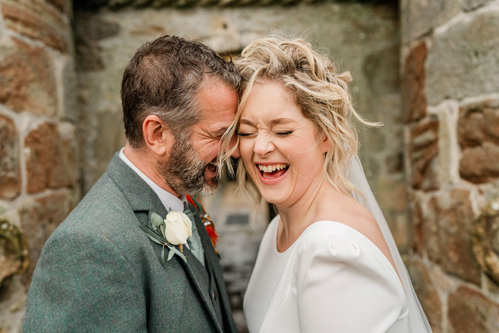 close up portrait image of bride in white wedding dress and veil and groom in grey suit jacket with a white buttonhole flower lean their foreheads into each other as they laugh