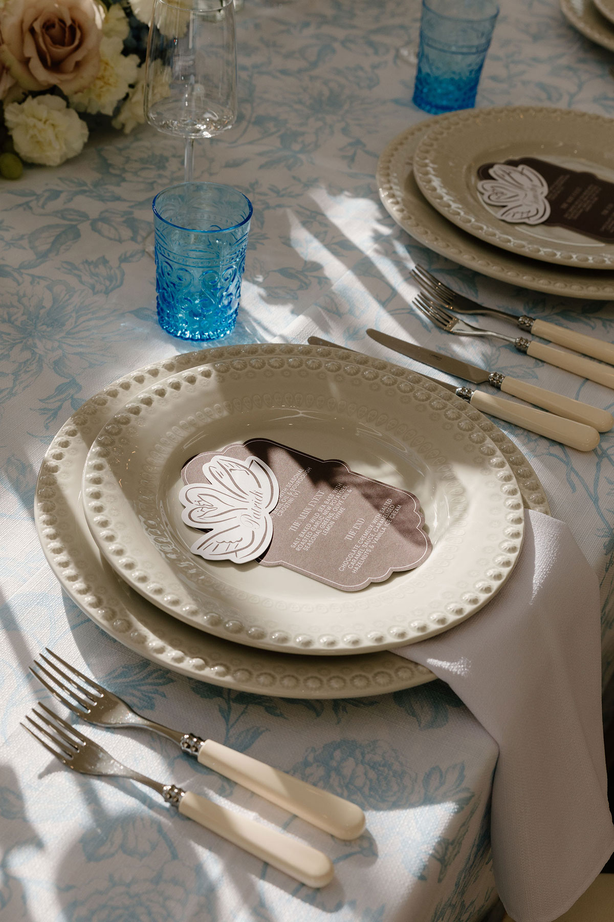 Place setting with cream plates, blue glassware and printed menu on a floral tablecloth