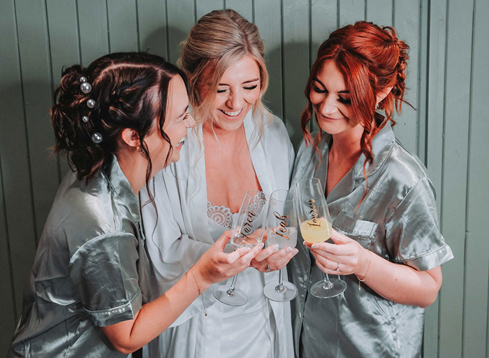 a bride and two bridesmaids wearing satin robes clinking personalised champagne flutes together