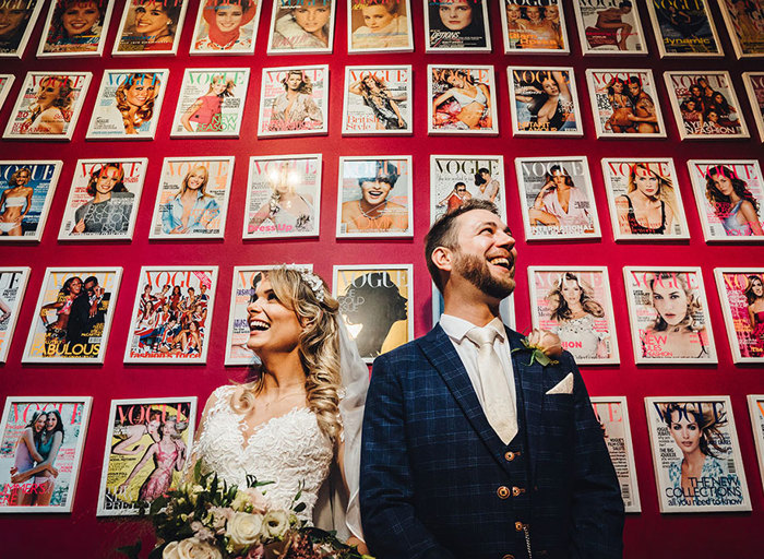 Bride and groom stand in front of a large red wall covered in framed Vogue magazine covers
