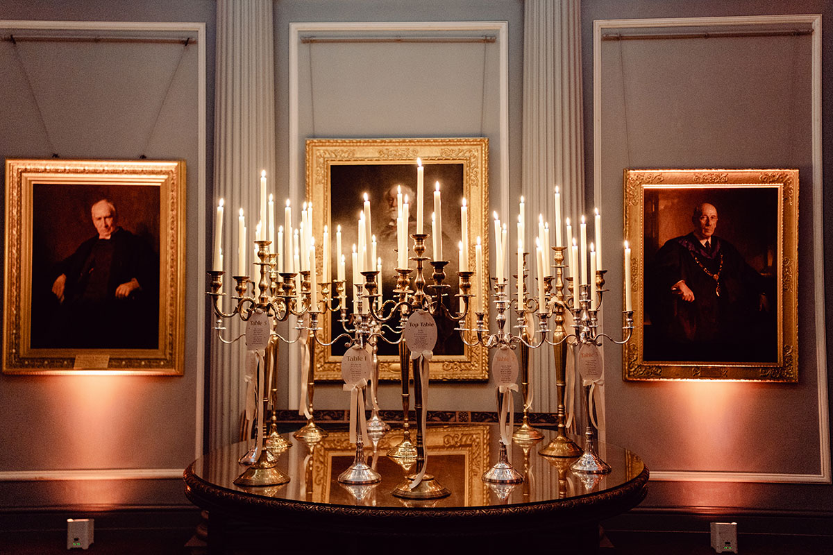 Large candelabras filled with tall candles displayed on a polished table inside the Signet Library, surrounded by traditional framed portraits