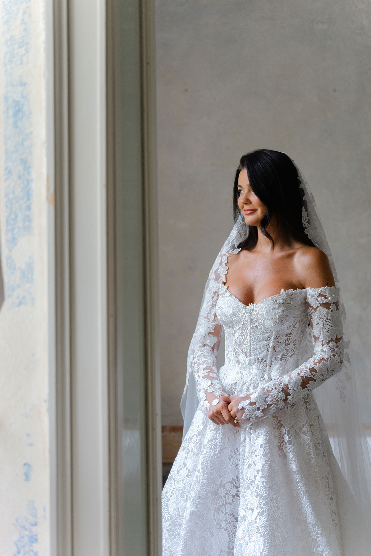 Bride in lace off-the-shoulder wedding dress standing by a window at Villa Regina Teodolinda, Lake Como