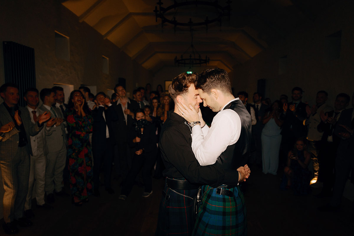 Two grooms share an emotional dance moment in matching Gunn tartan kilts as guests look on during their wedding reception at Newhall Estate
