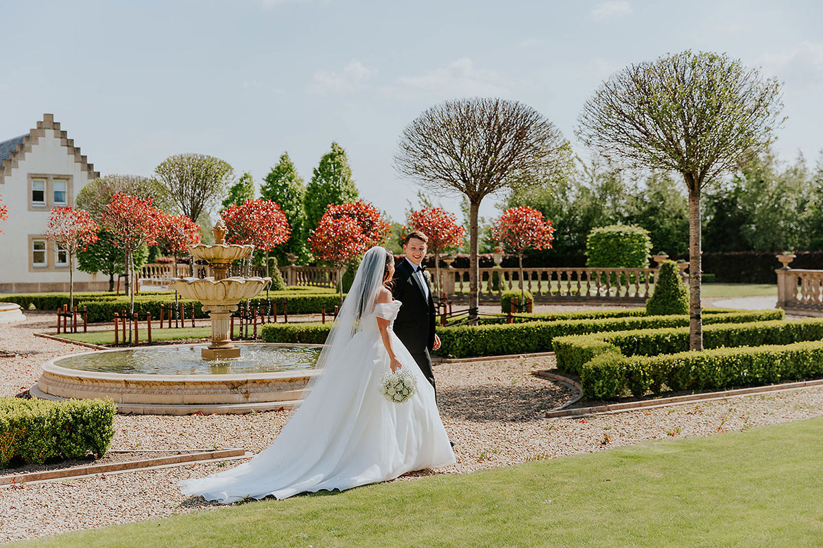 bride and groom walk through ingliston gardens with red trees on a sunny day