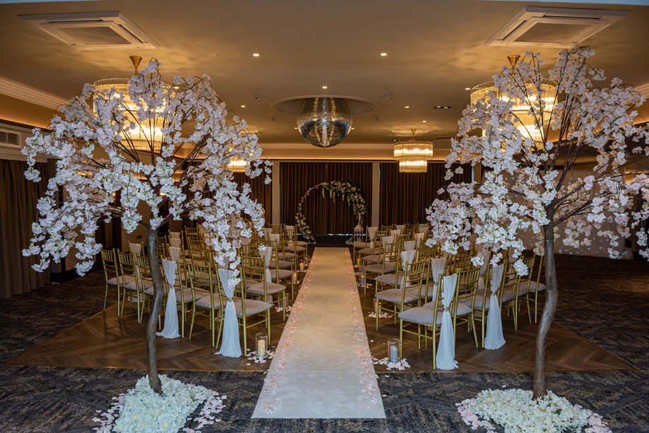 a white aisle runner is covered in pale pink and white flower petals with gold chairs with pink cushions and white drapes either side