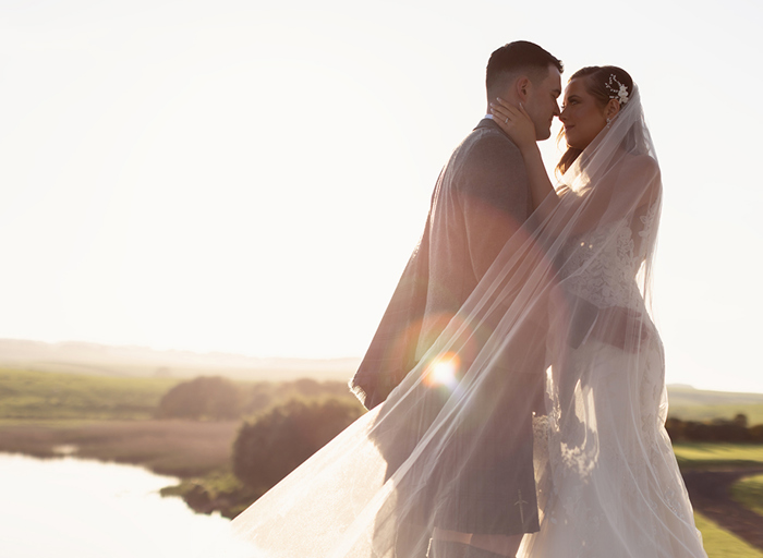 a bride and groom standing nose to nose basking in a golden sunset with green landscape and lake in background