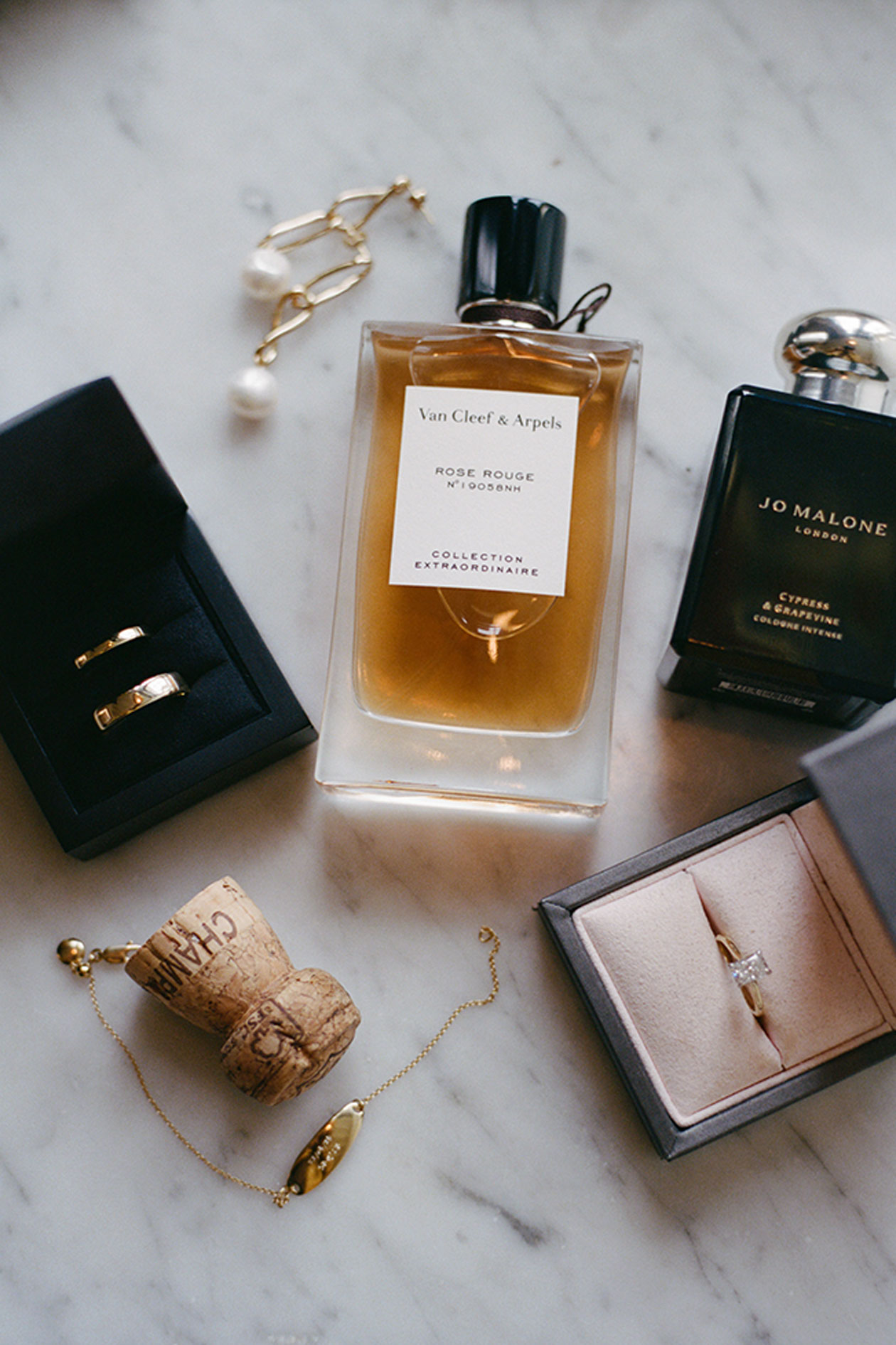 Alt: Wedding details flat lay with rings perfume earrings and champagne cork on marble surface