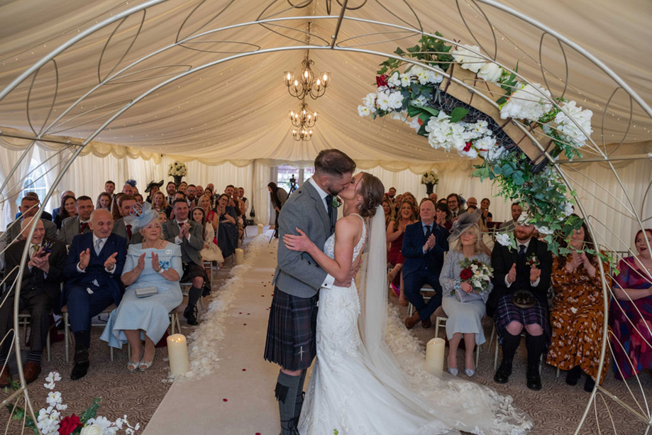 A Bride And Groom Kissing In Front Of Clapping Guests In A Marquee