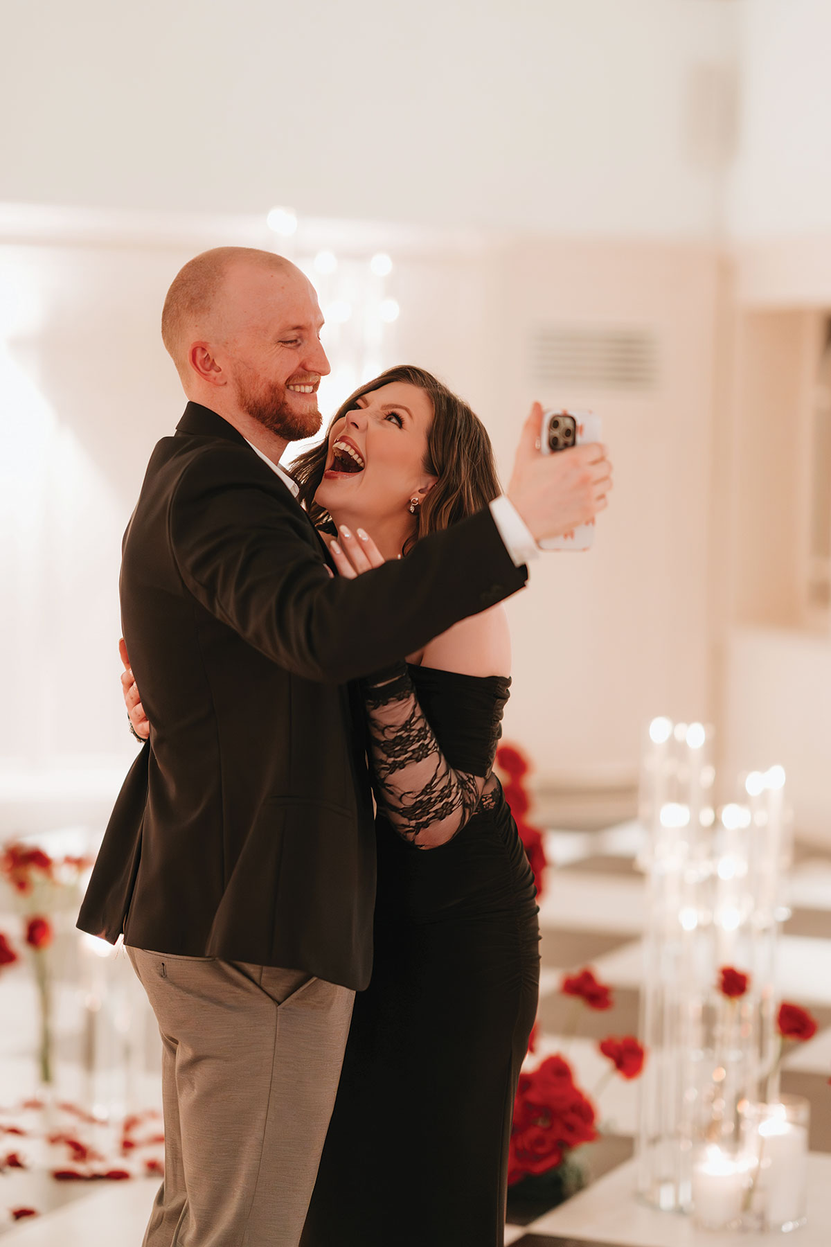 Couple taking a selfie after a surprise engagement proposal surrounded by candles and roses