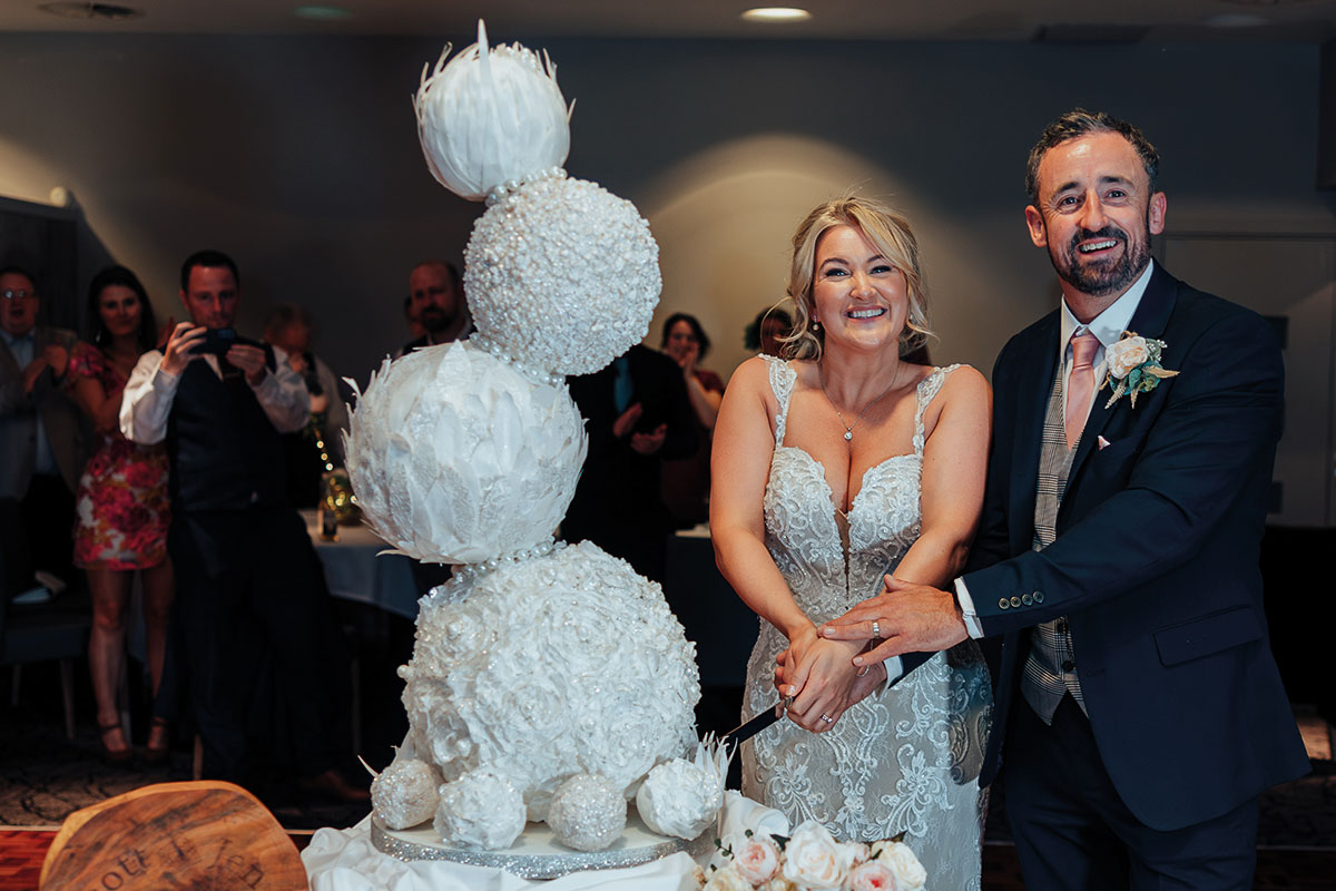 Bride and groom cutting a sculptural white wedding cake made from textured spherical tiers adorned with feathers and pearls, captured by Lloyd Stevenson Photography