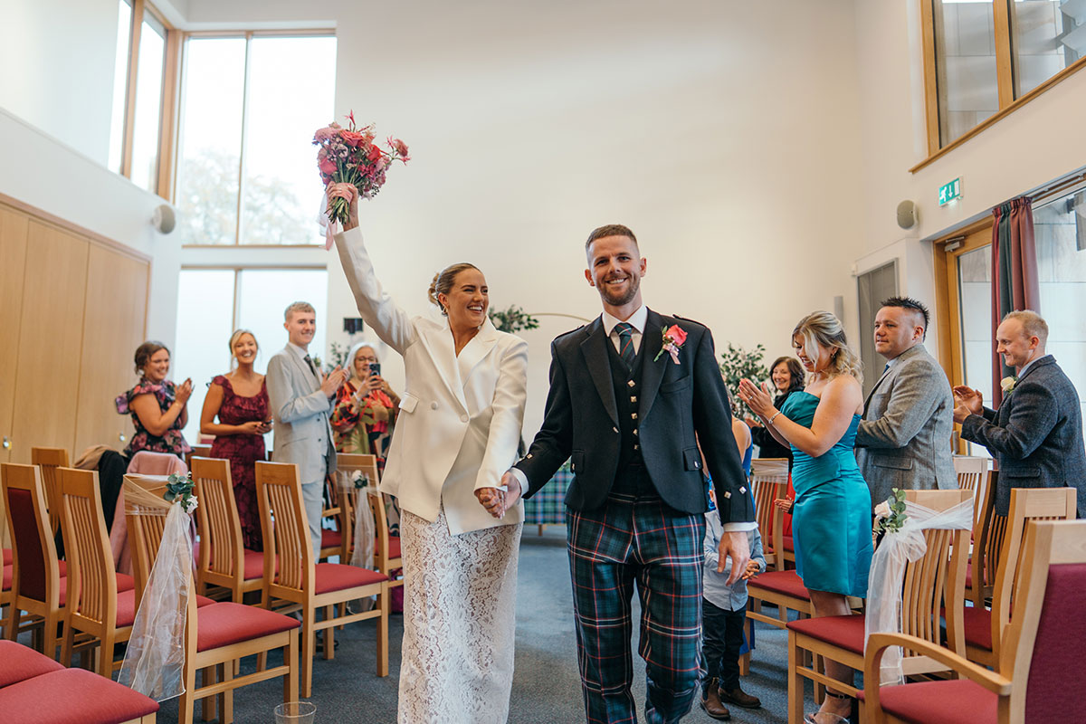 Newlywed couple walking back up the aisle hand in hand after their ceremony, with the bride raising her bouquet as guests applaud.
