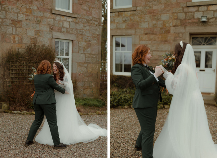 first look of two brides outside a Georgian-style building, one wearing a wedding dress, one wearing a dark green suit. They clasp hands and look elated