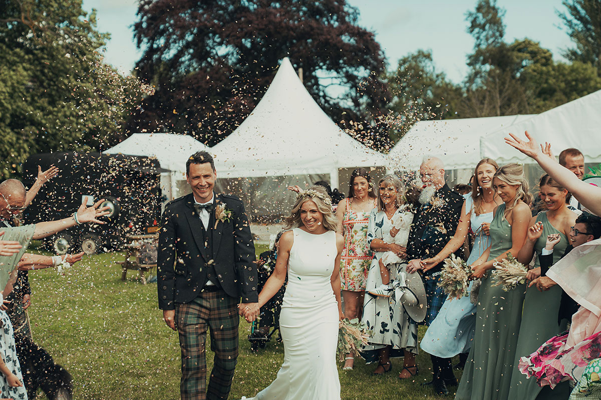 Newlyweds walk hand in hand through a shower of confetti outdoors, surrounded by cheering guests at a marquee wedding reception