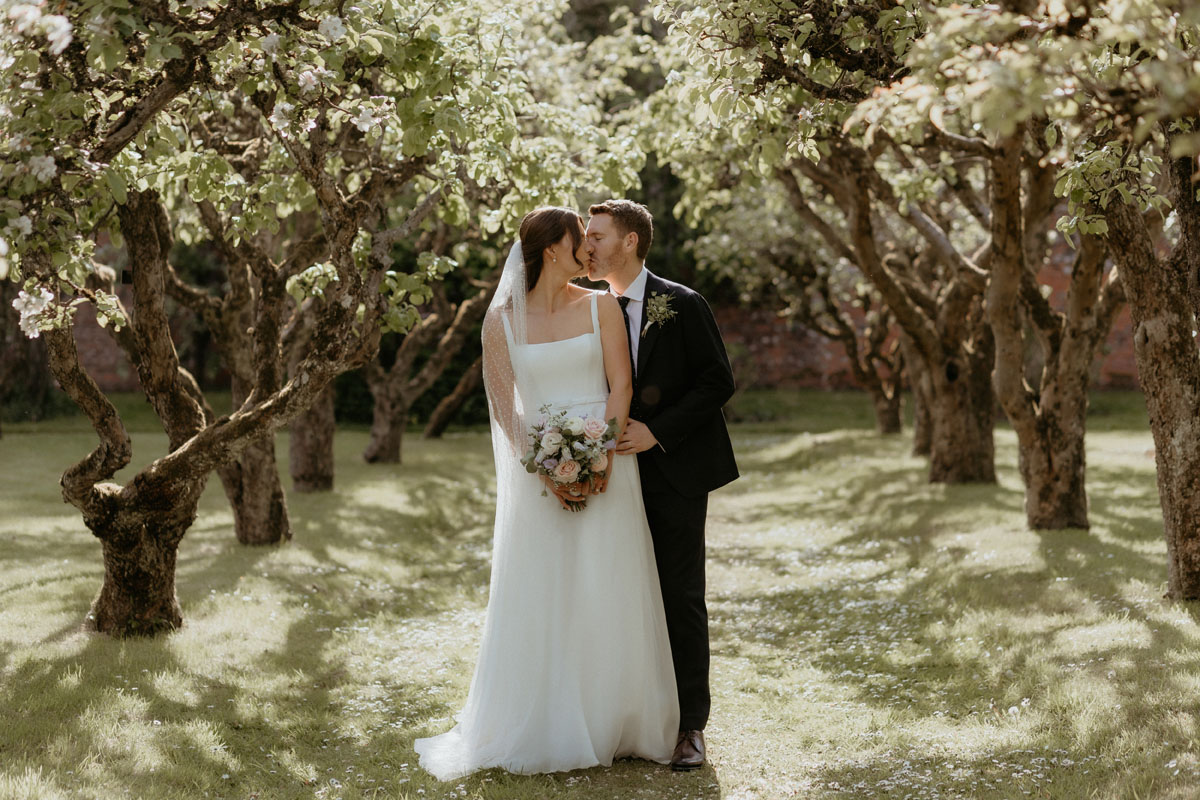 Bride and groom kissing beneath the trees in the orchard at The Byre at Inchyra in Perthshire.