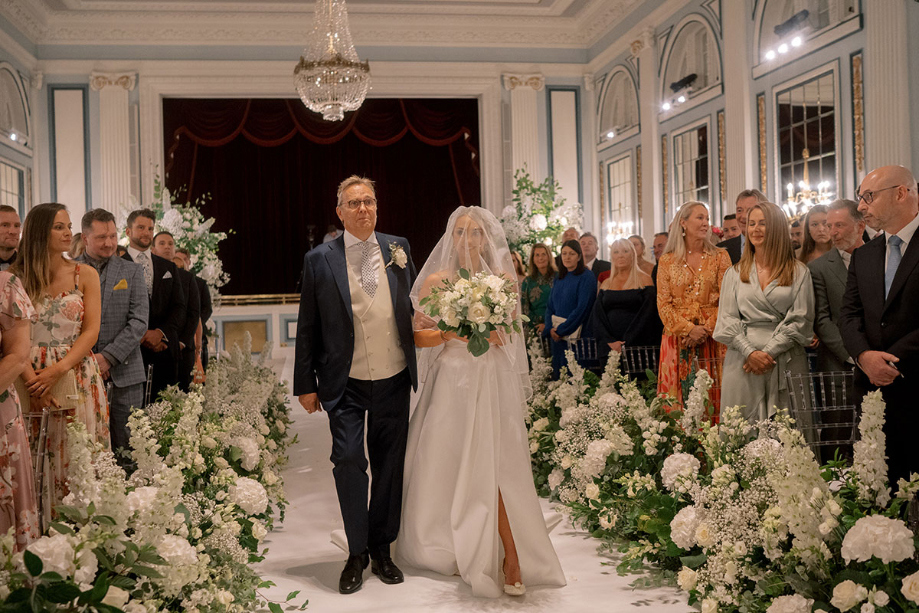 Bride and her father walk down the aisle that is lined with white flowers