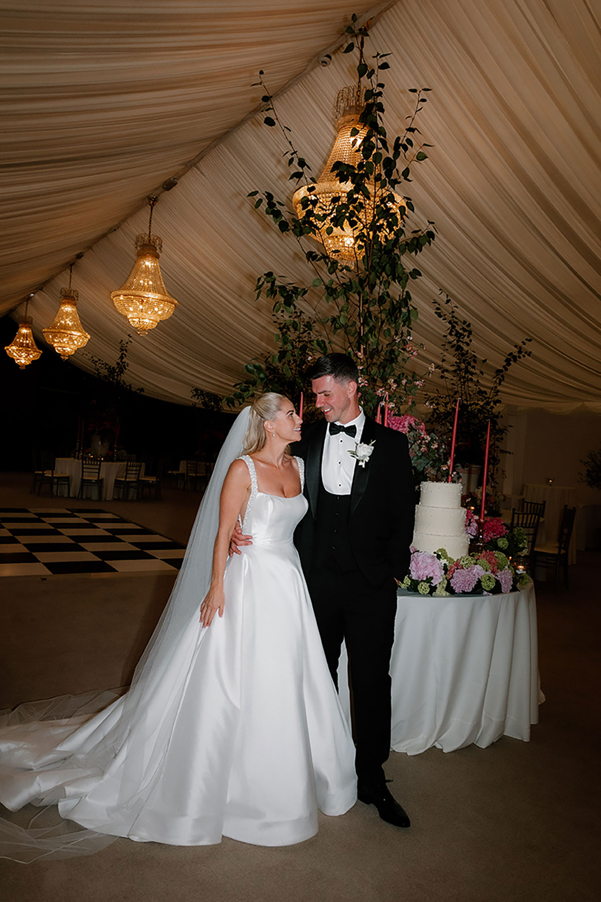 Bride and groom standing beside their wedding cake under chandeliers in the evening marquee at Mar Hall.