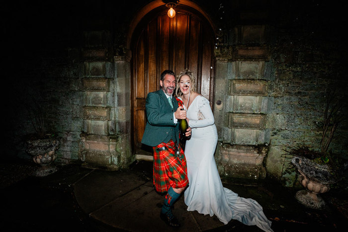 bride in white wedding dress and and groom in red and green tartan kilt stand outdoors in the dark, side by side, as they shake and spray a bottle of champagne in the air