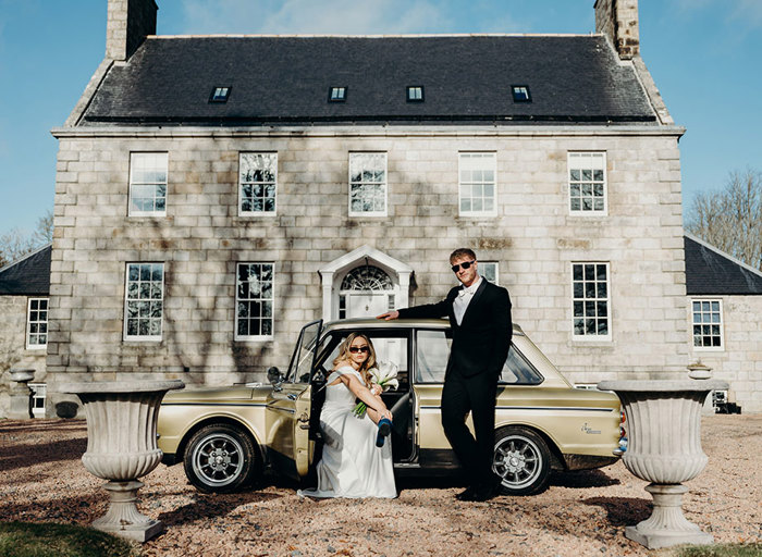 a bride and groom wearing sunglasses posing with a vintage gold car outside an elegant Georgian mansion