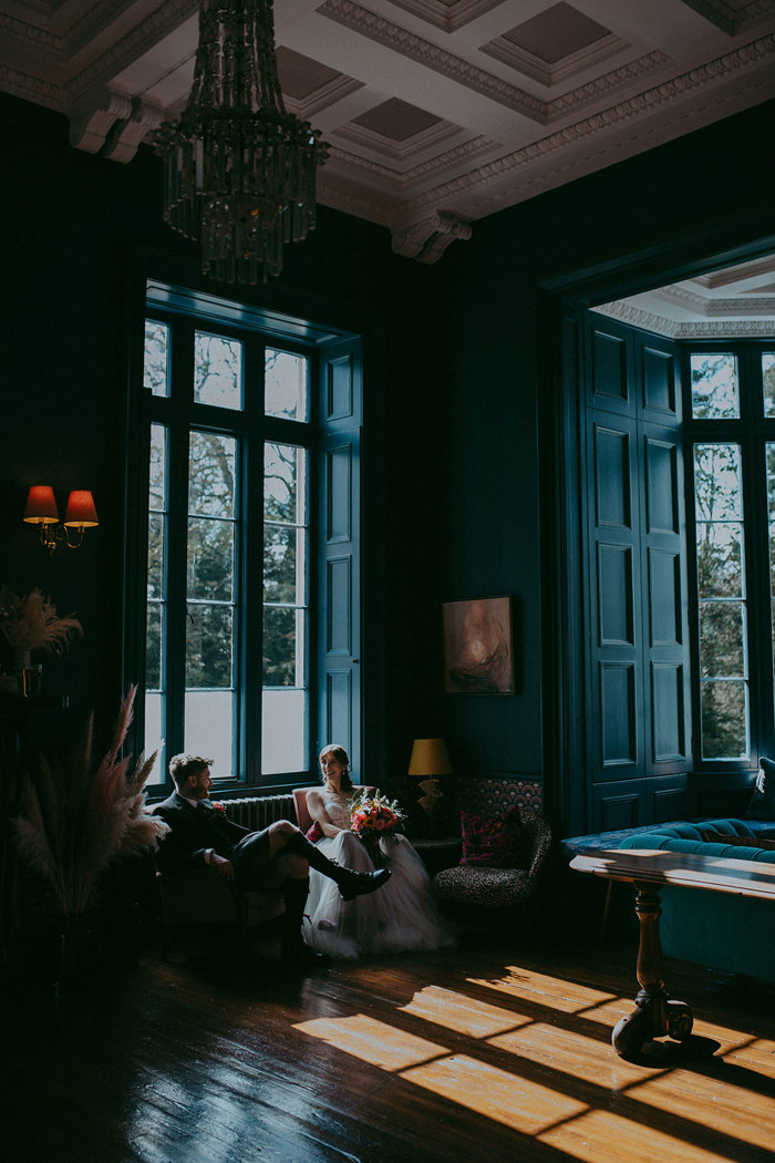 a bride and groom seated by a window in a dark teal room with sunlight streaming in onto a wooden floor at Netherbyres House