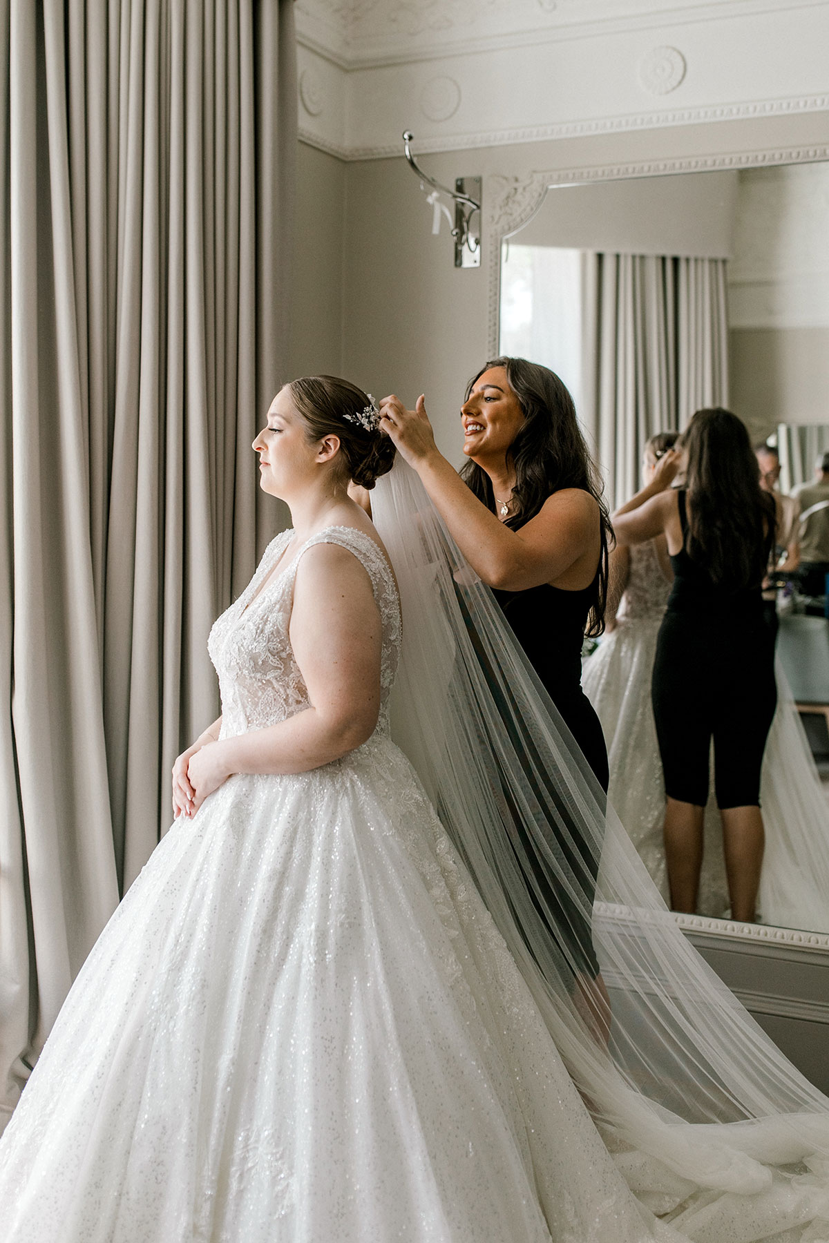 brunette woman in all black outfit smiles as she adjusts the veil of brunette bride in a large sparkly princess wedding dress ballgown