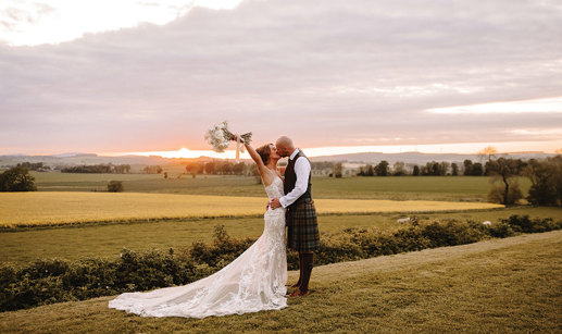 a bride wearing a lace dress and groom wearing a kilt kiss in a field of grass at sunset. The bride is holding her bouquet in the air