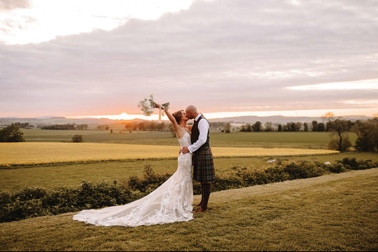 a bride wearing a lace dress and groom wearing a kilt kiss in a field of grass at sunset. The bride is holding her bouquet in the air