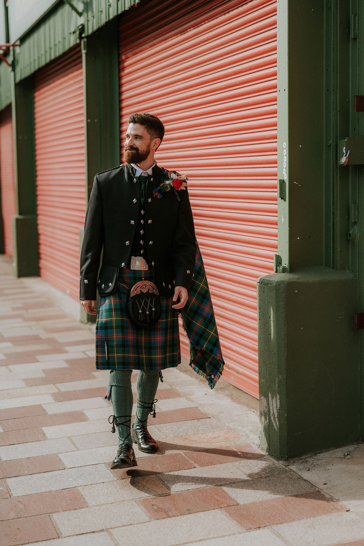 Groom in tartan kilt walking past red shutters at BAaD Glasgow wedding venue exterior