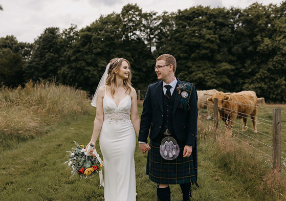 A bride and groom look at each other as they walk towards the camera along a verge of grass with a Highland Cow in a filed behind them
