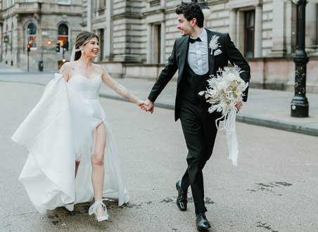 A bride and groom walk through a city holding hands 