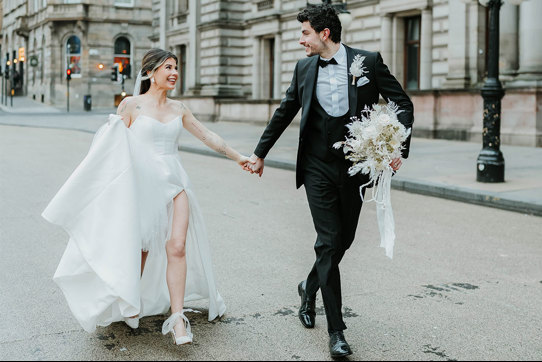 A bride and groom walk through a city holding hands 