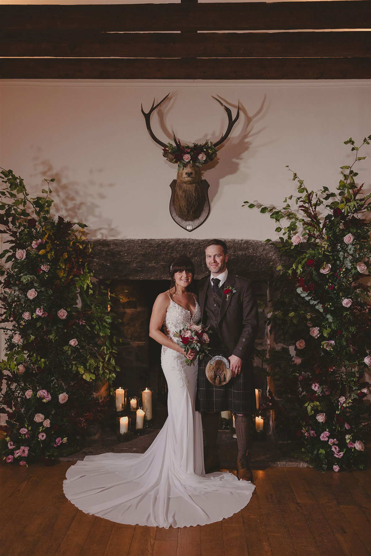 Bride in a white lace wedding dress and groom in a dark kilt outfit posing in front of Aswanley's stone fireplace, framed by large floral arrangements and candles beneath a stag's head